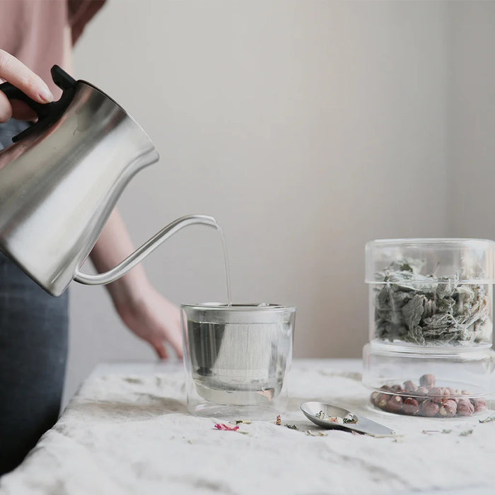 Person pouring tea from a silver kettle into a glass on a marble surface.