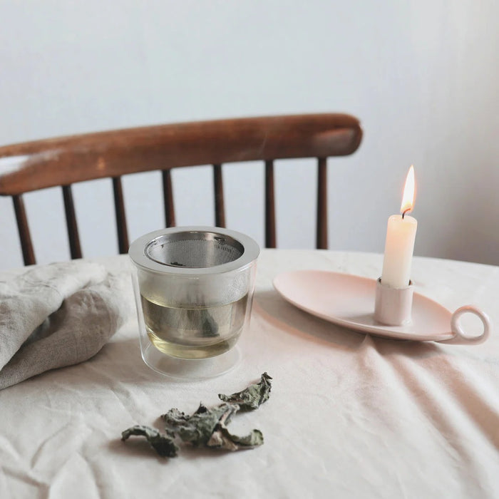 Clear glass of tea with a tea bag, dried tea leaves, and a lit candle on a wooden chair.